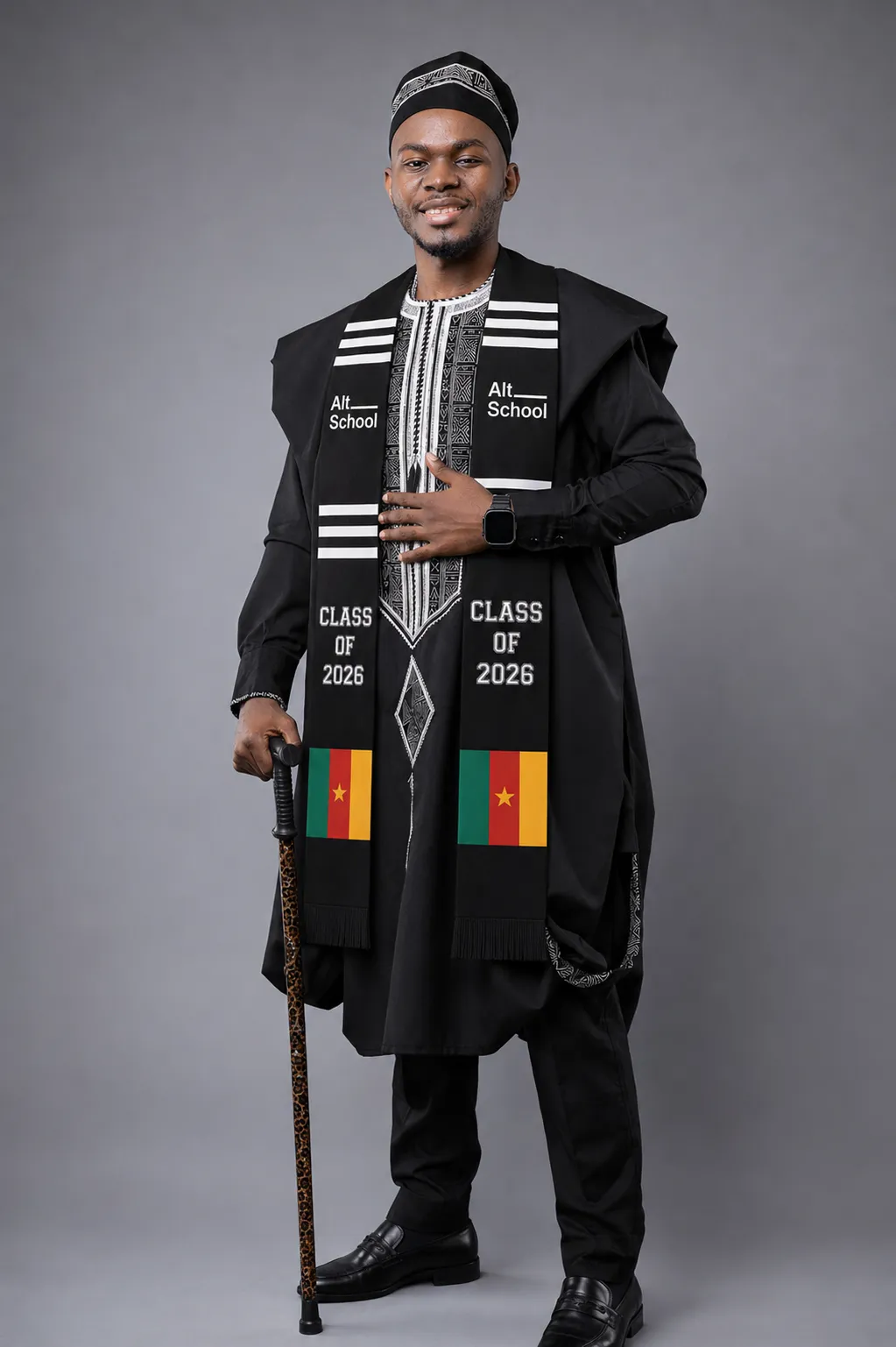 Michel Tegofack in a black traditional outfit and cap, wearing the AltSchool "Class of 2026" stole with Cameroon flags, cane in hand, on a grey backdrop.